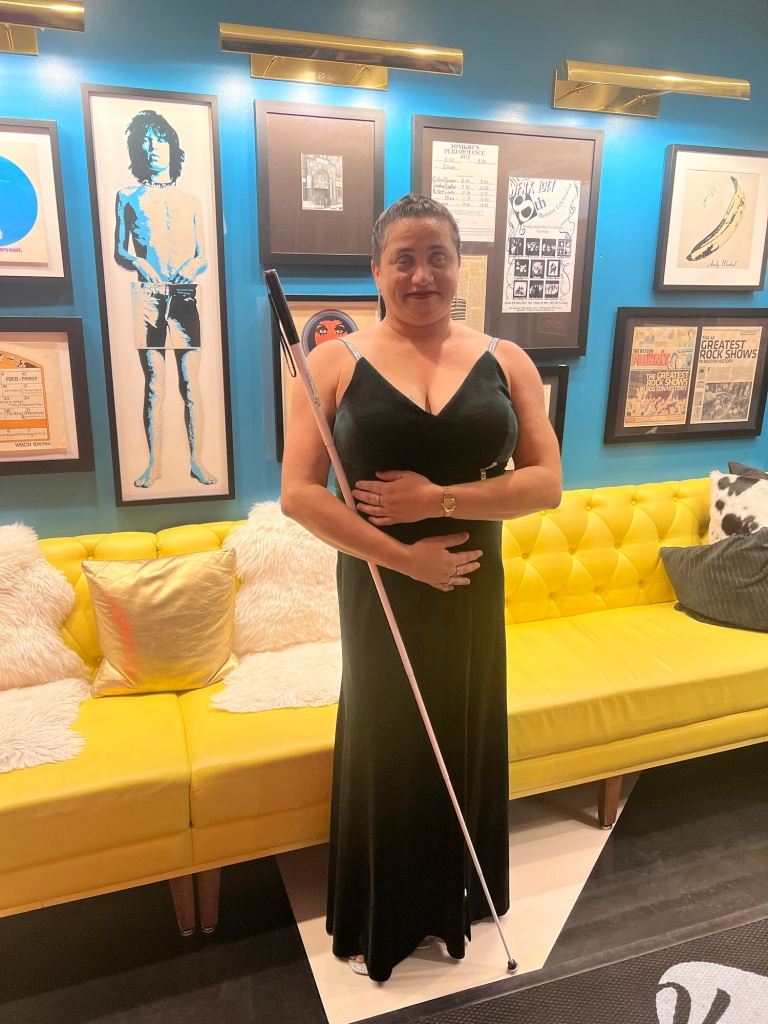 Lisamaria stands in a hotel lobby and she is wearing a velvet, green, evening dress. Her cane stands against her right shoulder. ￼
